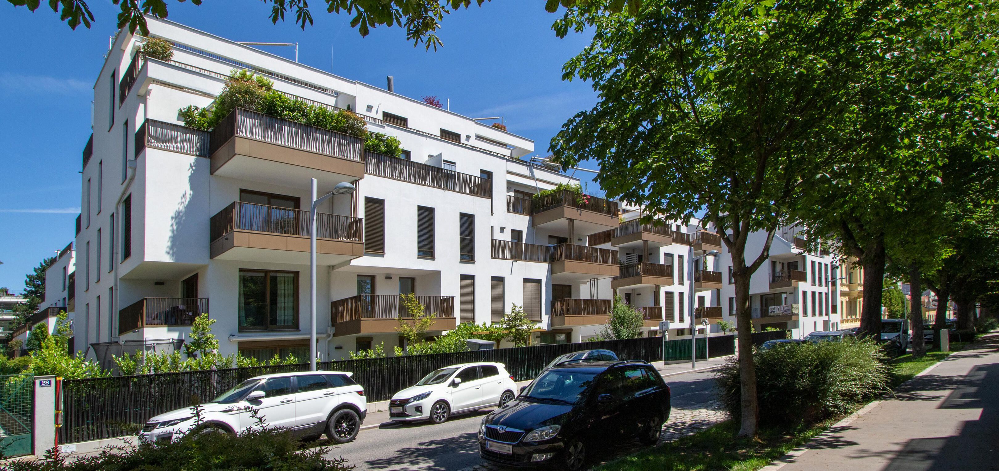 Blick von der Straße auf das Wohnbauprojekt mit schattigen Bäumen davor